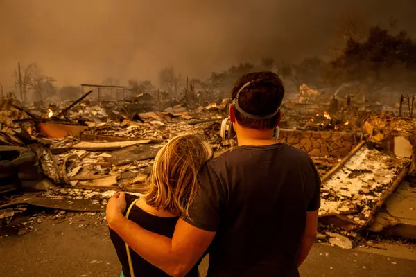 Two residents observing the aftermath of the Eaton Fire with charred trees and smoke in the background.